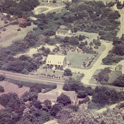 Old photograph of an overhead view of a house; the other houses in the photo are distant from the main house, separated by trees and a road winding through.