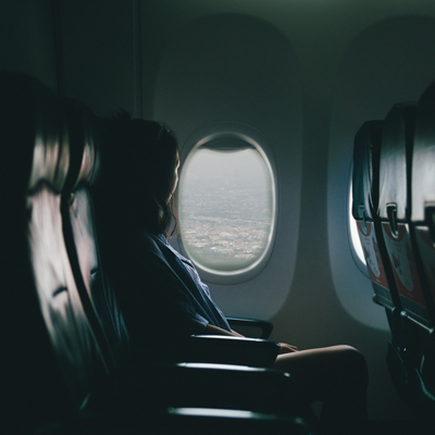 A woman sits by the window of a shadowed airplane. The window provides most of the light as she looks out it.