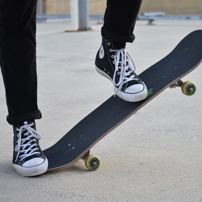 One foot (wearing black Converse) holds down one end of a skateboard, raising the other end up. The other foot rests gently a little more than halfway up the elevated skateboard.