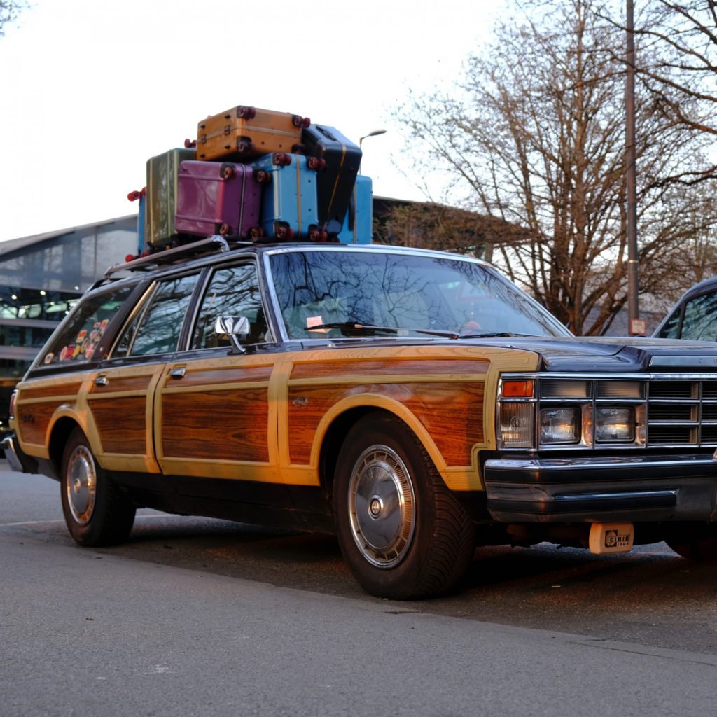 An Oldsmobile sits on the street with suitcases in various shapes and colors packed on top of it