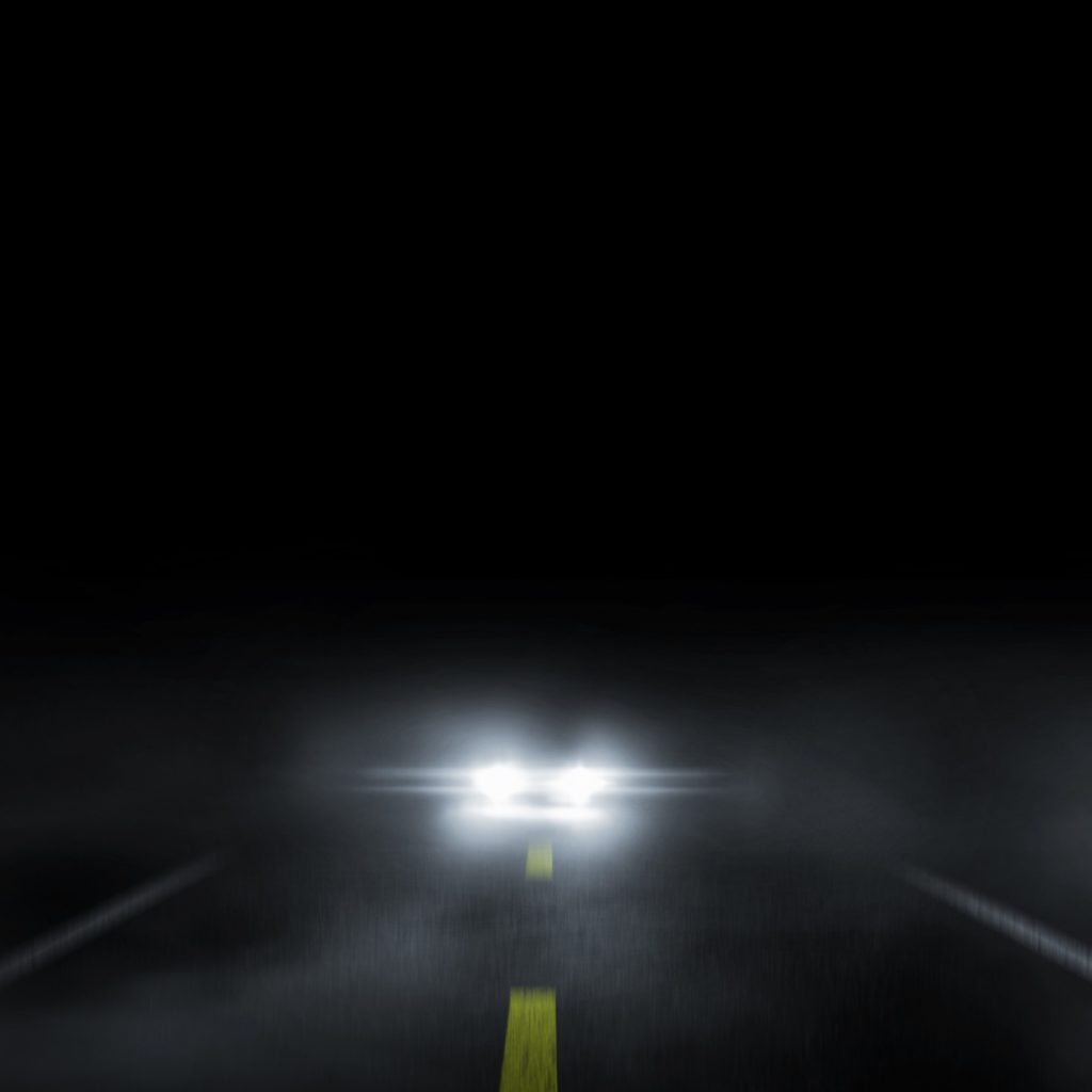 A car's headlights stare into the camera against a nighttime road and sky